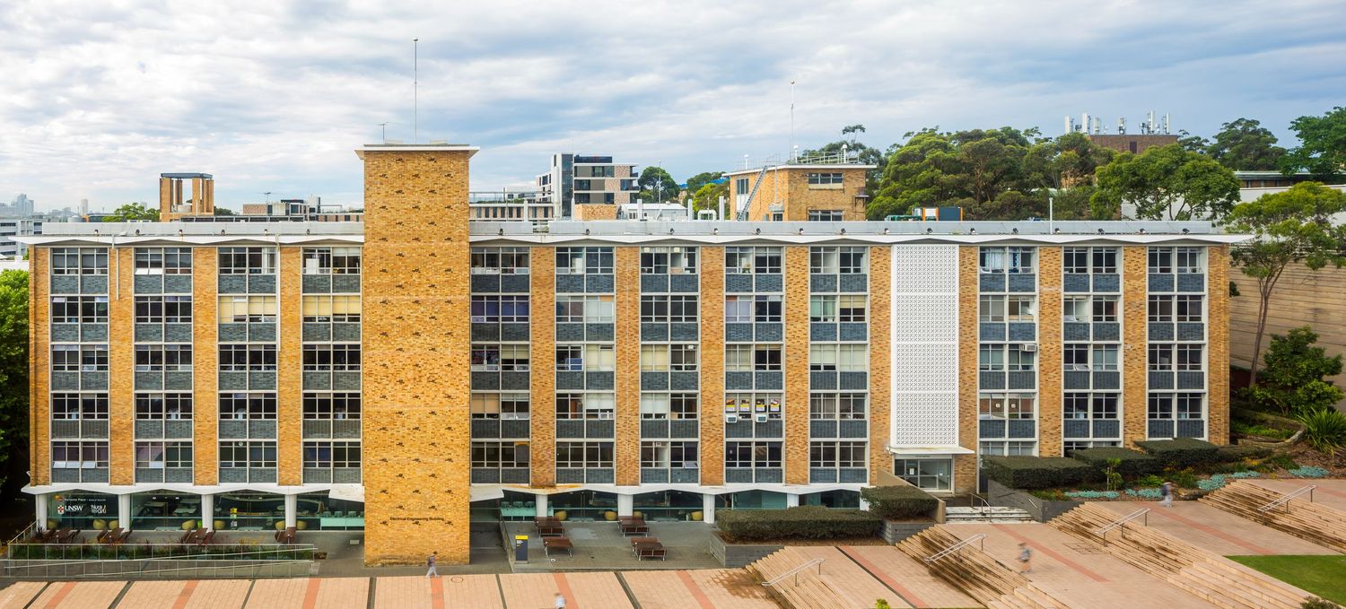 Mid-century apartment building with orange brick walls, balconies and a central stair tower beside trees.