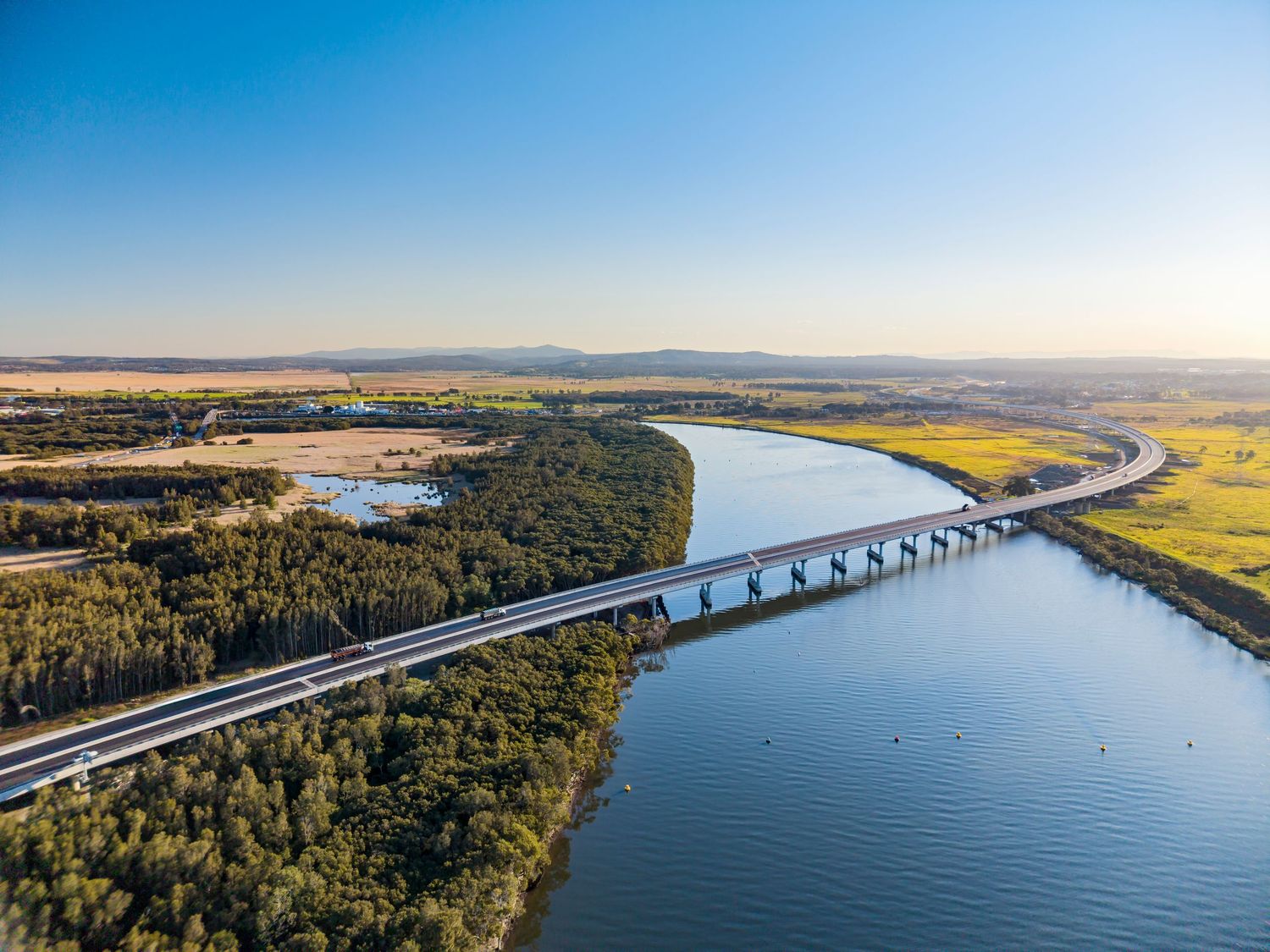 Aerial view of a long highway bridge crossing a wide river through green bushland and farmland at golden hour.