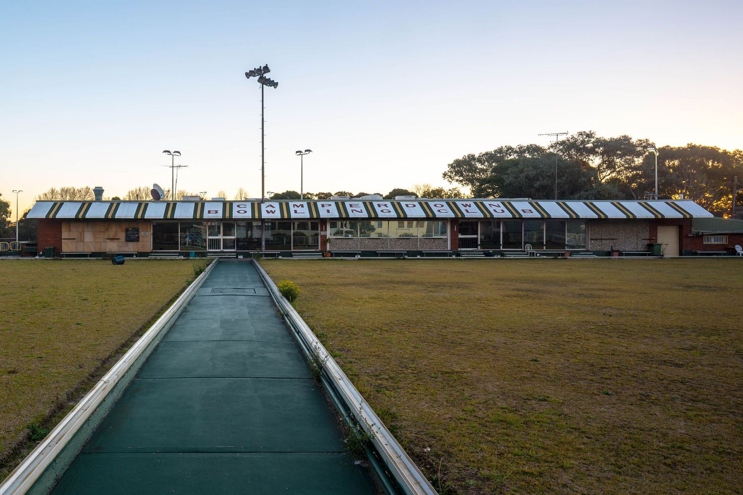Long green walkway leads across a grassy lawn toward a low brick pavilion with a sawtooth roof at dusk.