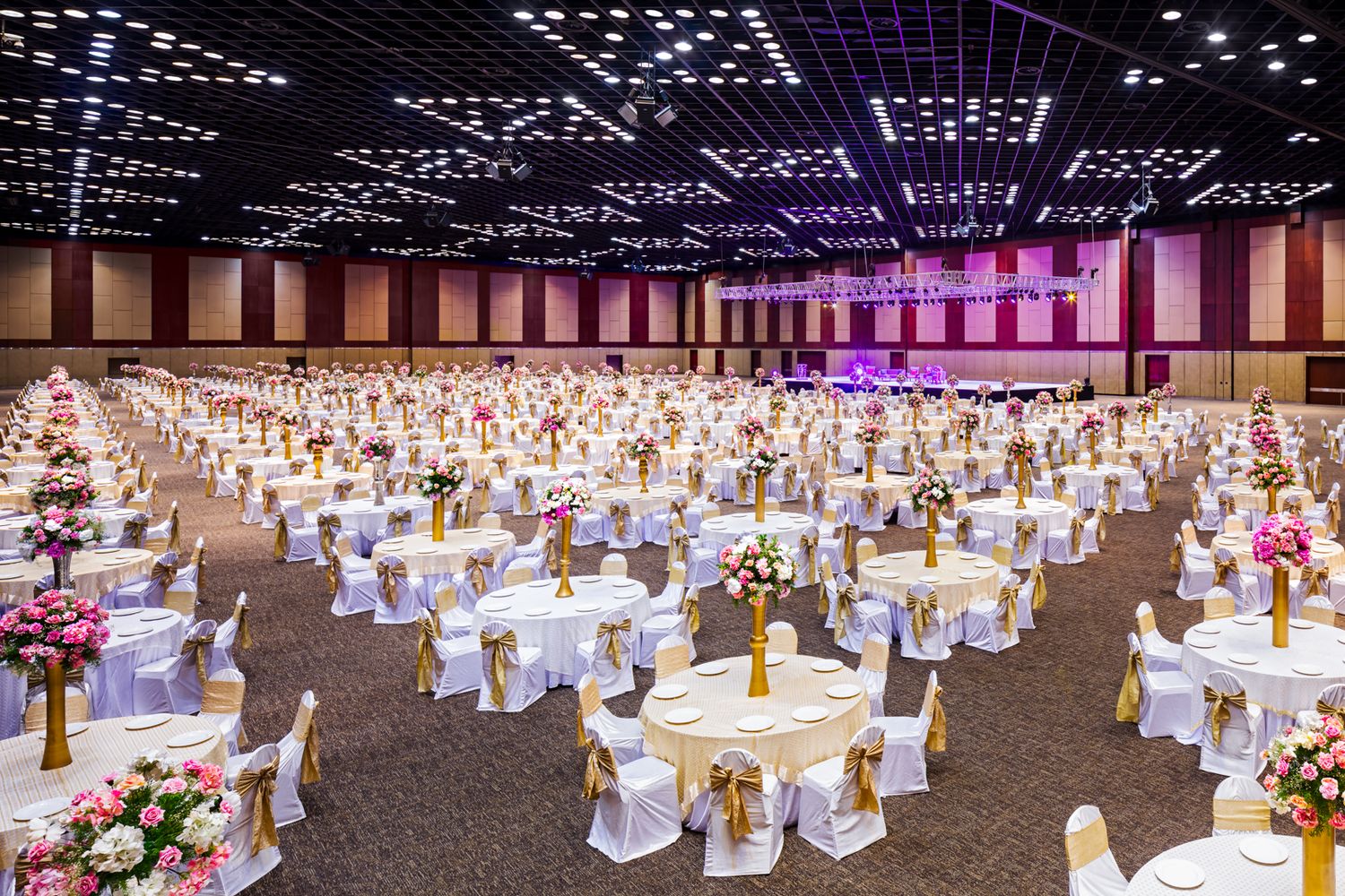 Spacious banquet hall set with round tables, white linens, gold ribbon chairs and pink floral centerpieces for a wedding or gala event.