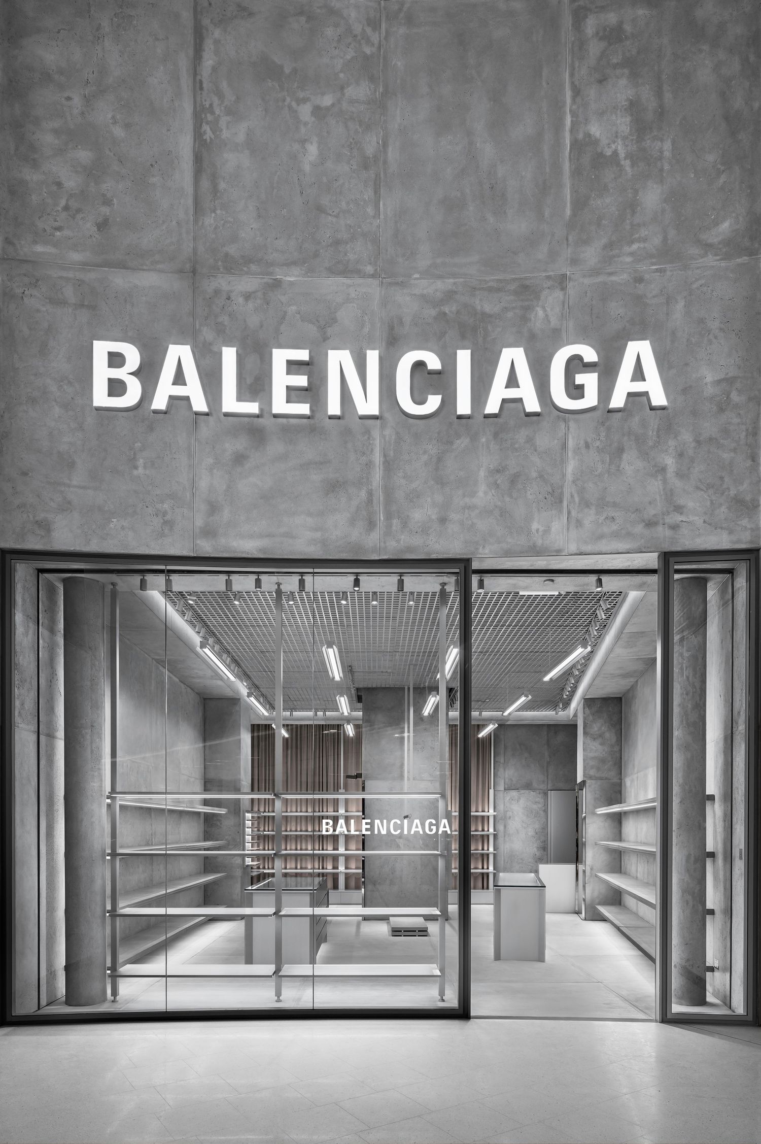 Balenciaga boutique entrance with white lettering on a raw concrete facade and a minimalist glass-fronted interior with display shelves.