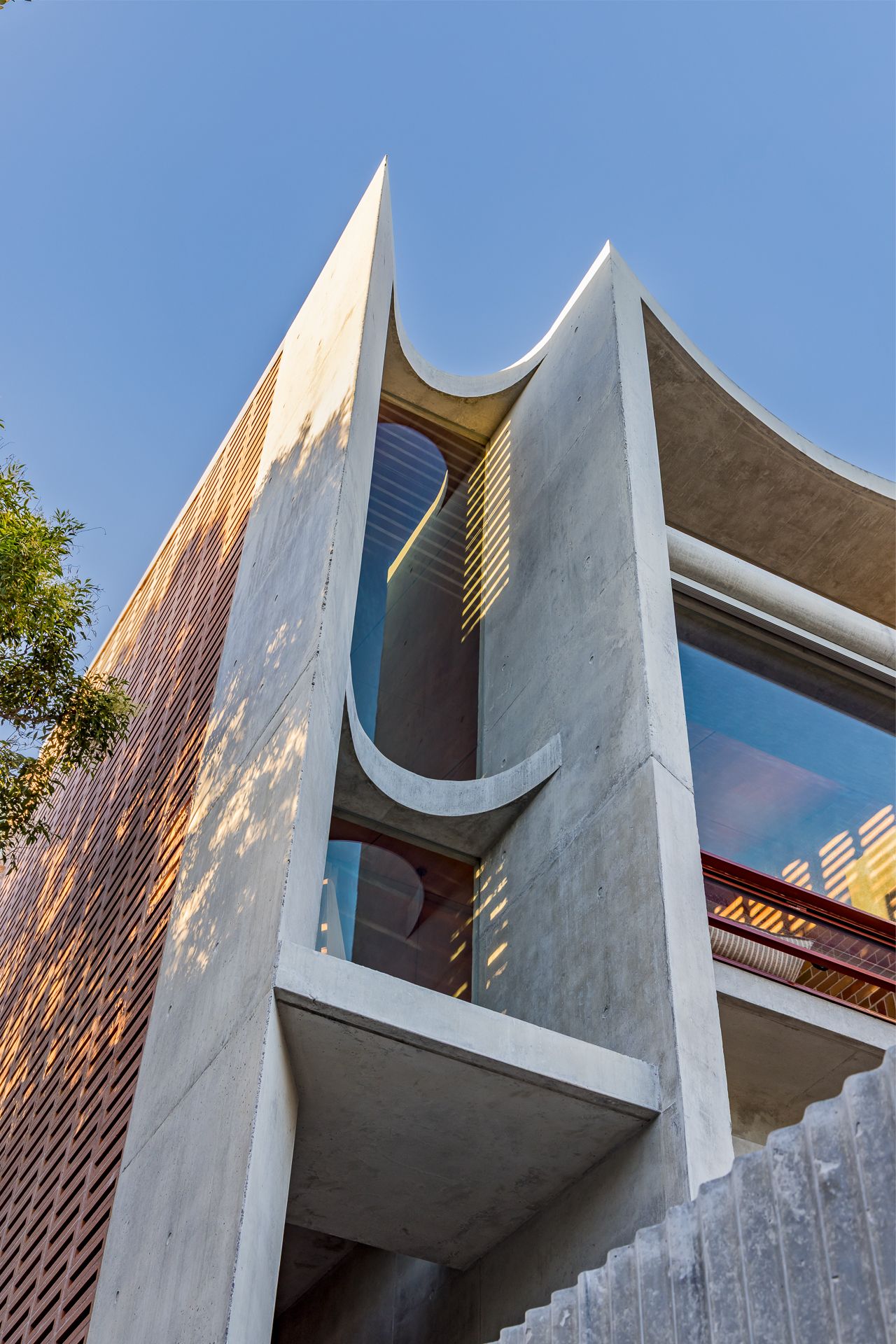 Sculptural concrete facade with sweeping curved cutouts, glass windows and a brick screen against a clear blue sky.