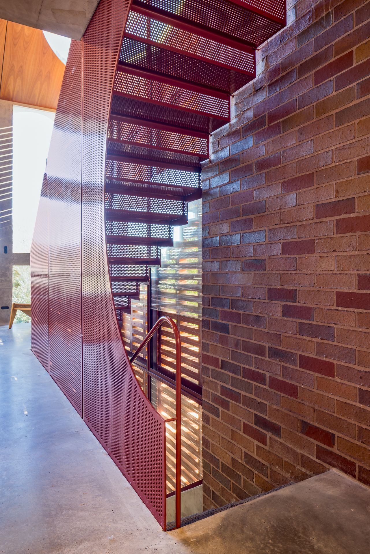 Red perforated metal staircase curving beside a textured brick wall, with sunlight filtering through slatted screens.
