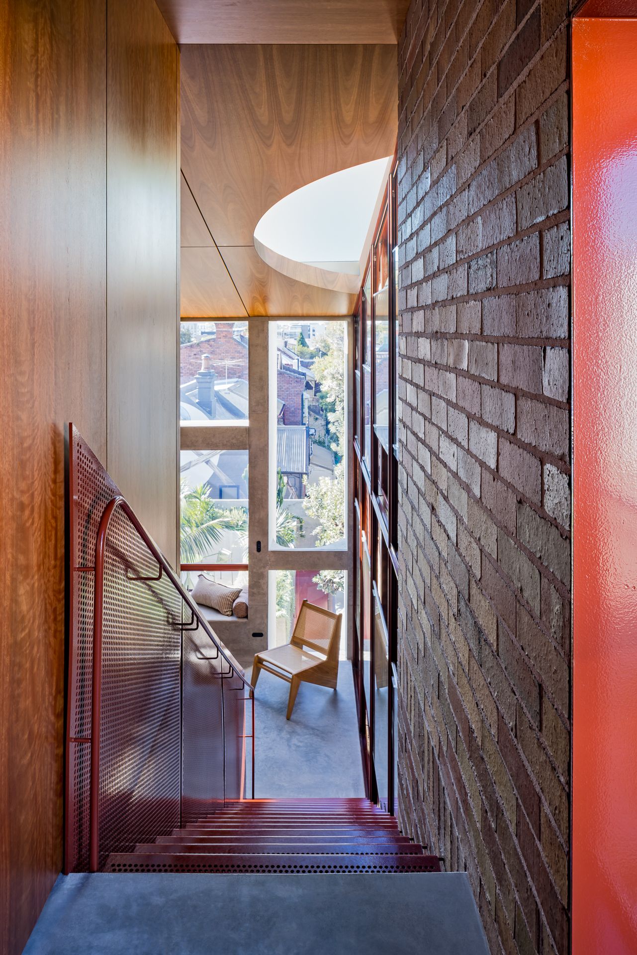 Stairwell with a red perforated metal balustrade, exposed brick wall, timber paneling, a skylight, and a wooden chair by a tall window.