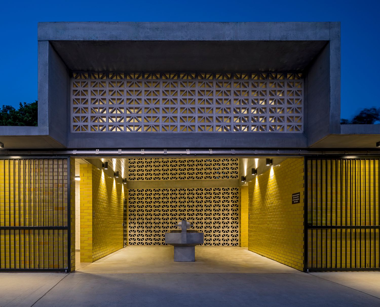 Modern concrete building entrance at dusk with decorative breeze block screens, glowing yellow walls, and a stone fountain in the courtyard.