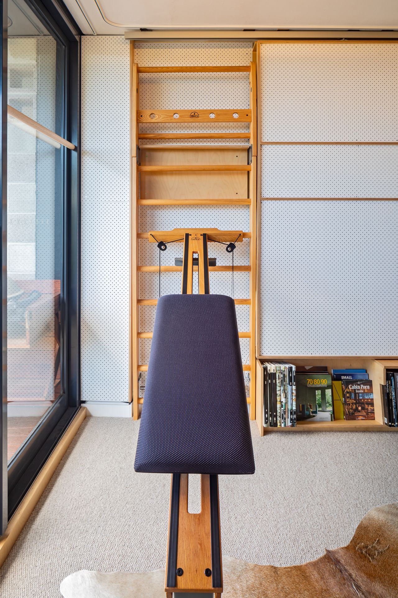 Padded exercise bench facing a wooden wall bars frame in a bright home gym with carpet, a window and a bookshelf.