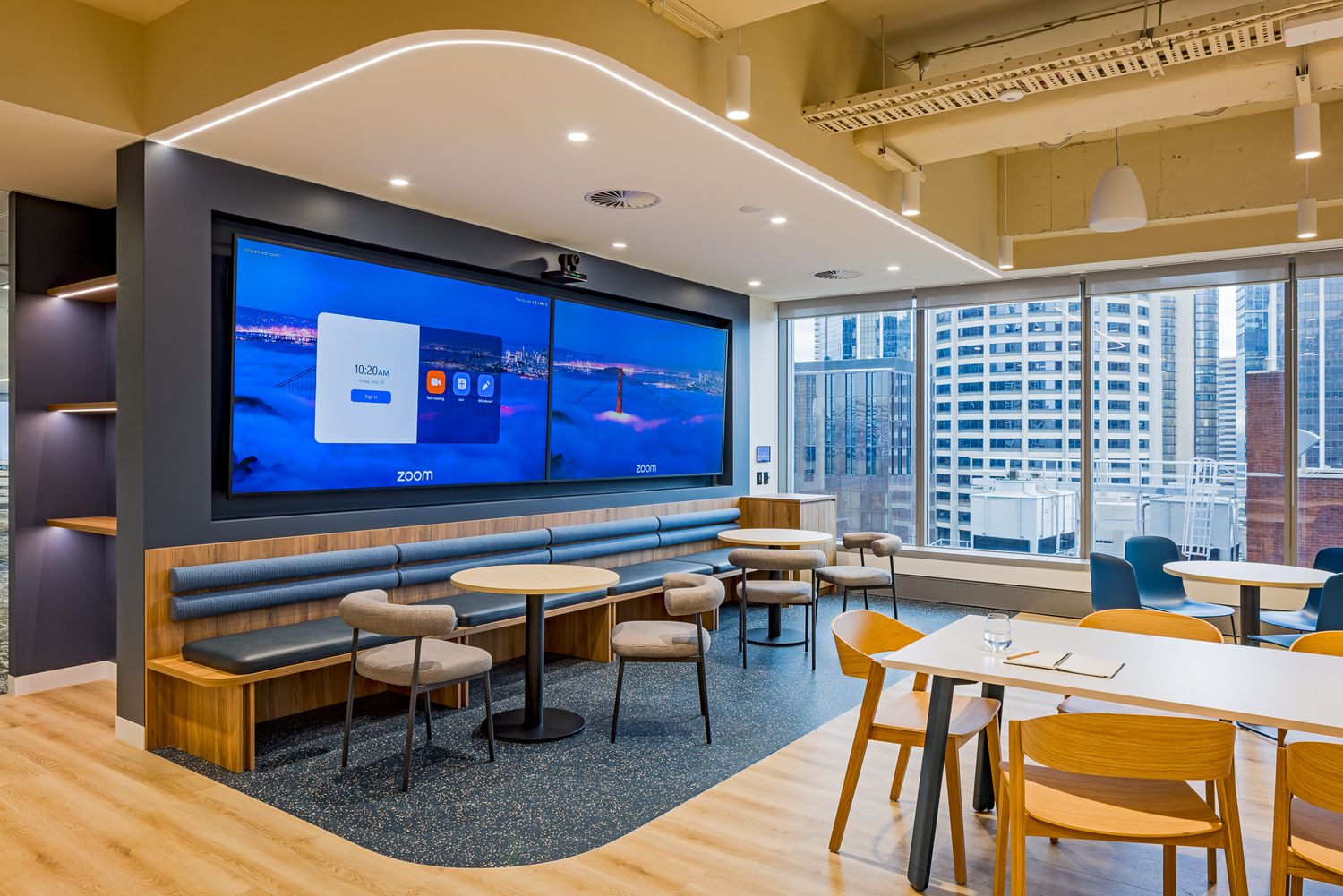Cozy office breakout area with built-in bench seating, round tables, a large video display and floor-to-ceiling city skyline windows.