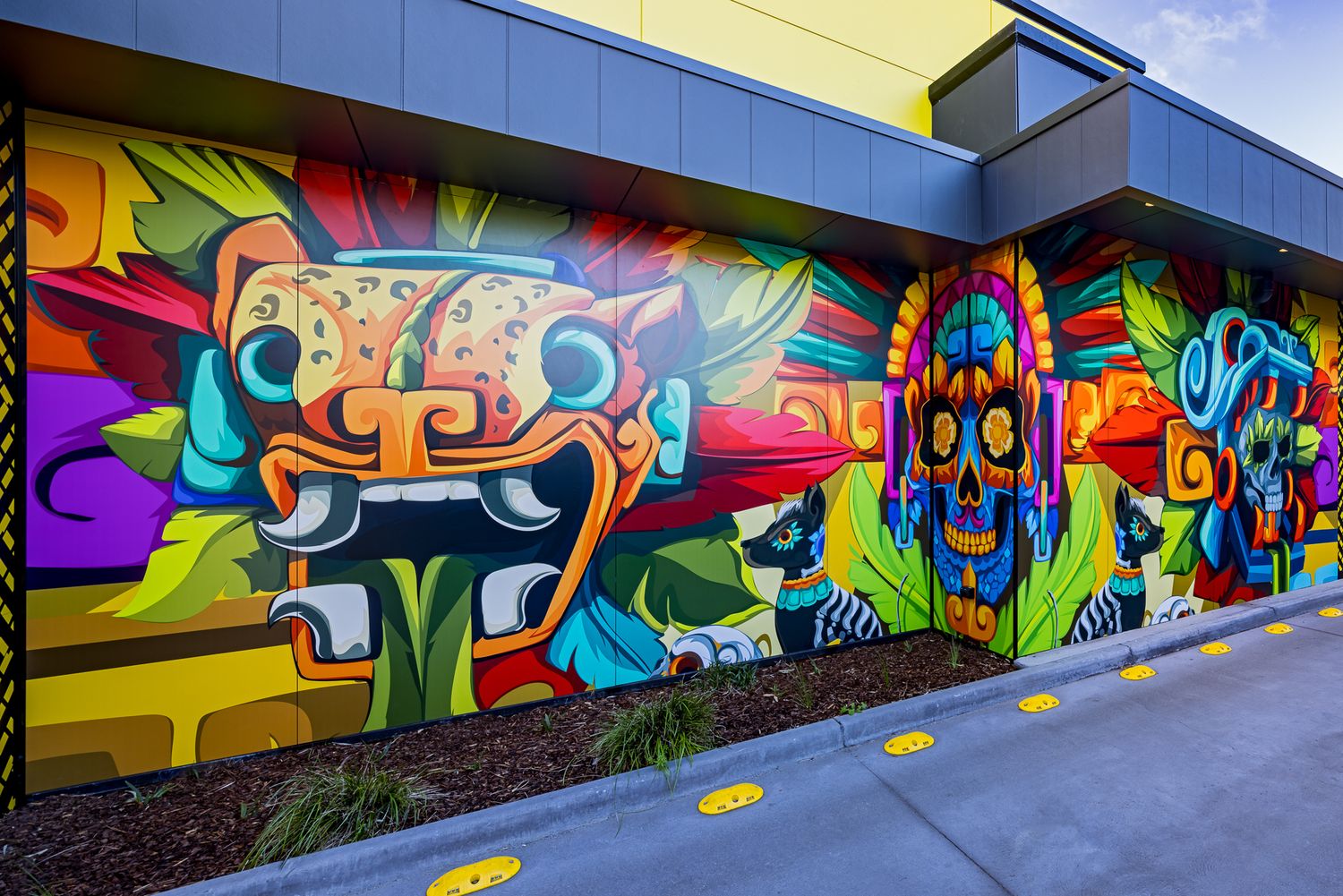 Vibrant street art mural on a restaurant wall featuring a jaguar head and skull in bright Aztec-inspired colors with yellow floor markers.