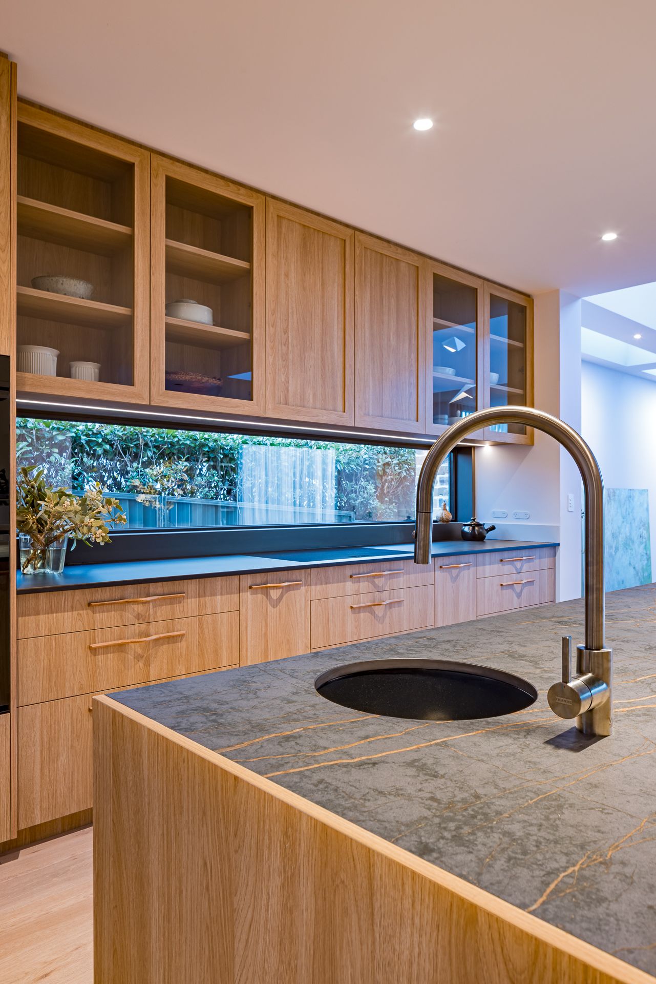 Close-up of a timber kitchen with glass-front cabinets, a stone island with veined top, round sink and curved brushed steel tap.
