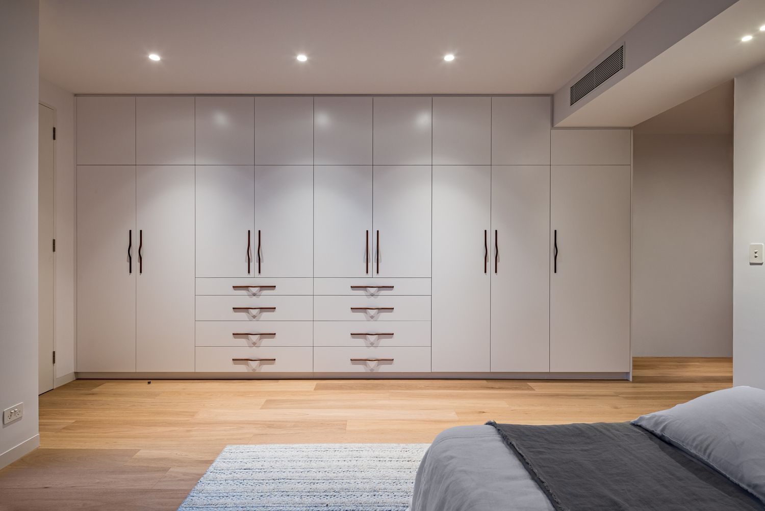 Bedroom with floor-to-ceiling white built-in wardrobes featuring slim black handles, recessed lighting, timber floors and a soft rug.