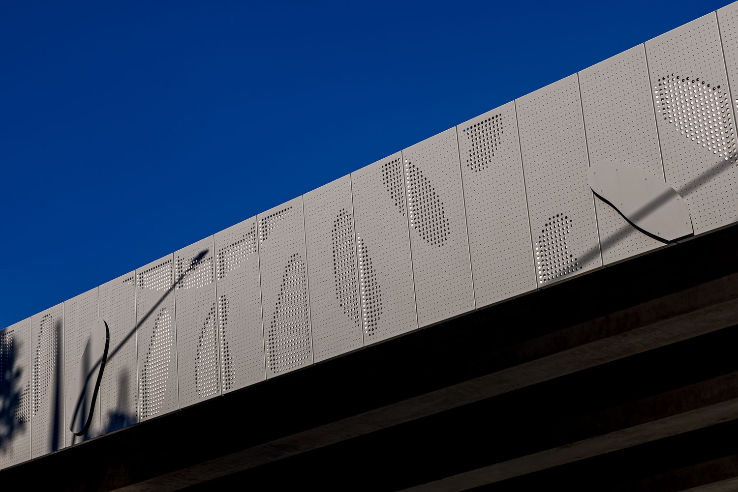 Close-up of perforated metal cladding panels with organic leaf-shaped cutouts along the edge of an elevated bridge against a deep blue sky.