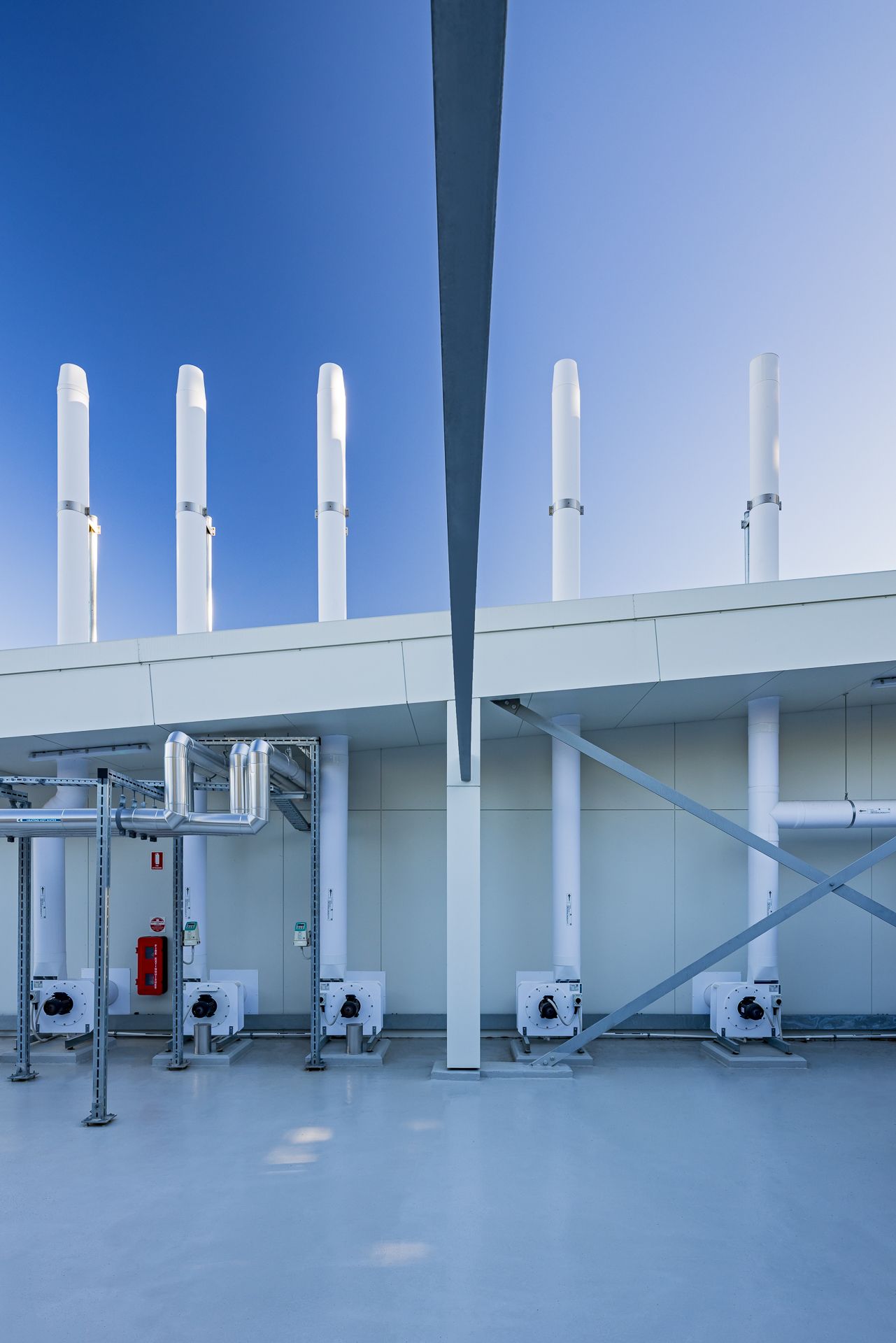 Row of white exhaust stacks above ventilation units and steel pipework on a building rooftop under a deep blue sky.