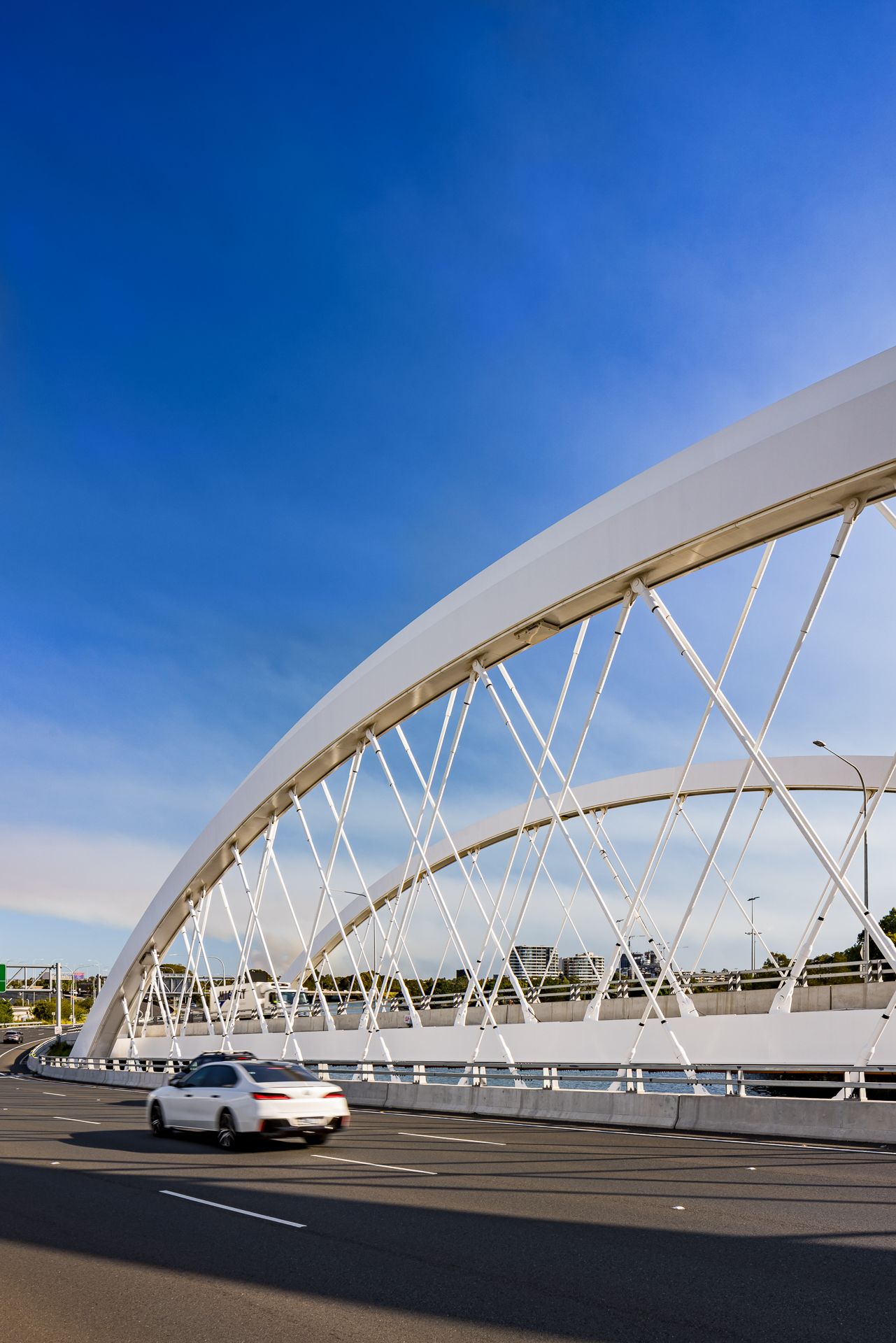 White sedan driving past the tall steel lattice arches of a modern bridge against a vivid blue sky with wispy clouds.