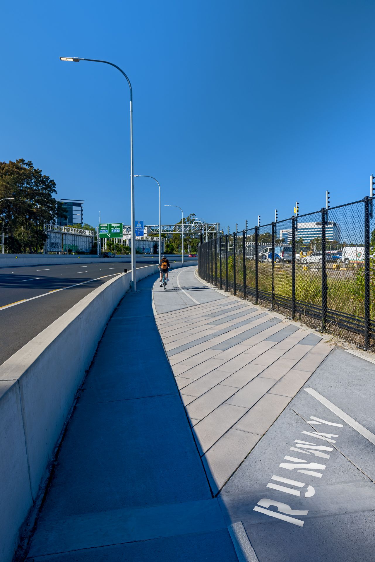 Dedicated cycleway beside a motorway with a cyclist riding along a fenced path marked with painted pavement lettering under blue sky.
