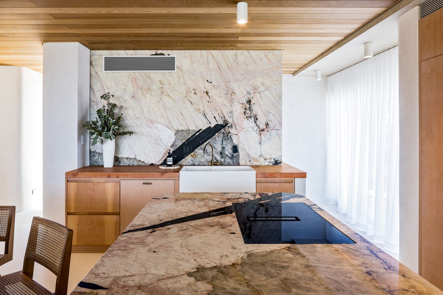 Contemporary kitchen with natural stone island and cooktop, timber cabinetry, farmhouse sink and a pale marble feature wall.