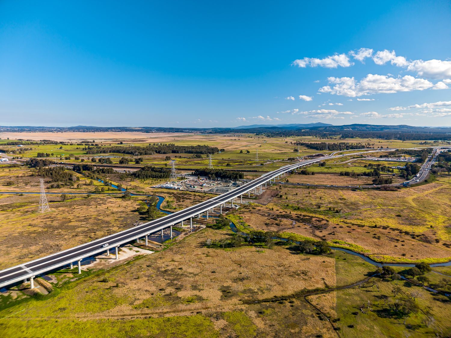 Aerial view of a highway viaduct crossing farmland and wetlands with distant mountains under a bright blue sky with clouds.