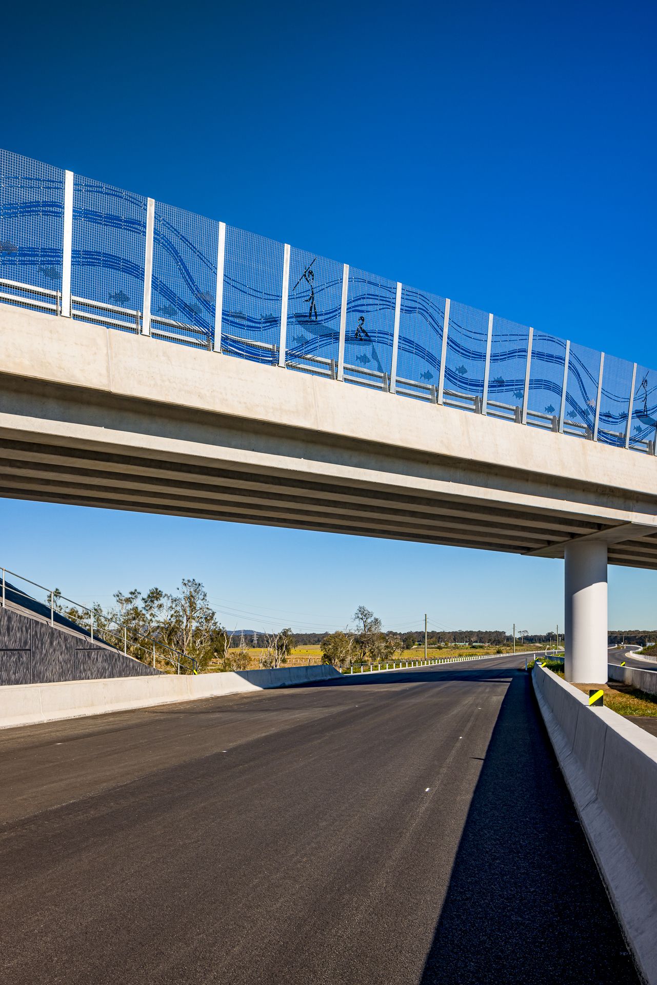 Highway overpass with patterned blue noise wall panels, supported by a round concrete pier above freshly paved road.