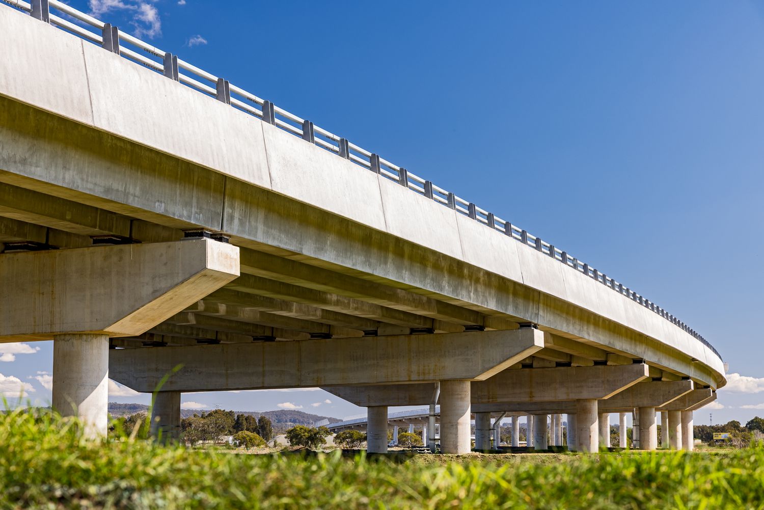 Low-angle view of a concrete viaduct supported by sturdy pillars above green grass and distant hills.