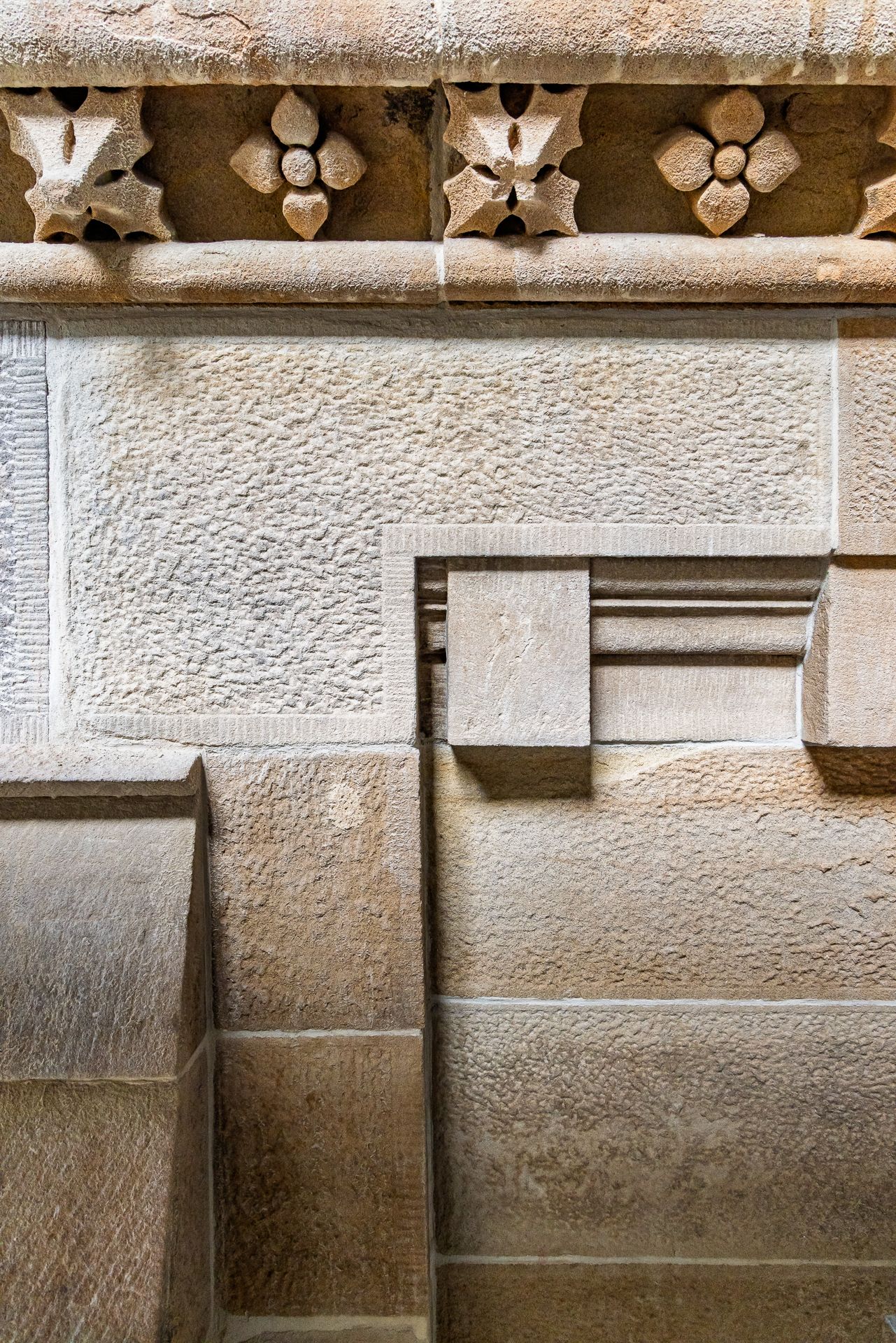 Detail of carved stone flower ornaments along a cornice above tooled sandstone blocks with a recessed stepped panel.