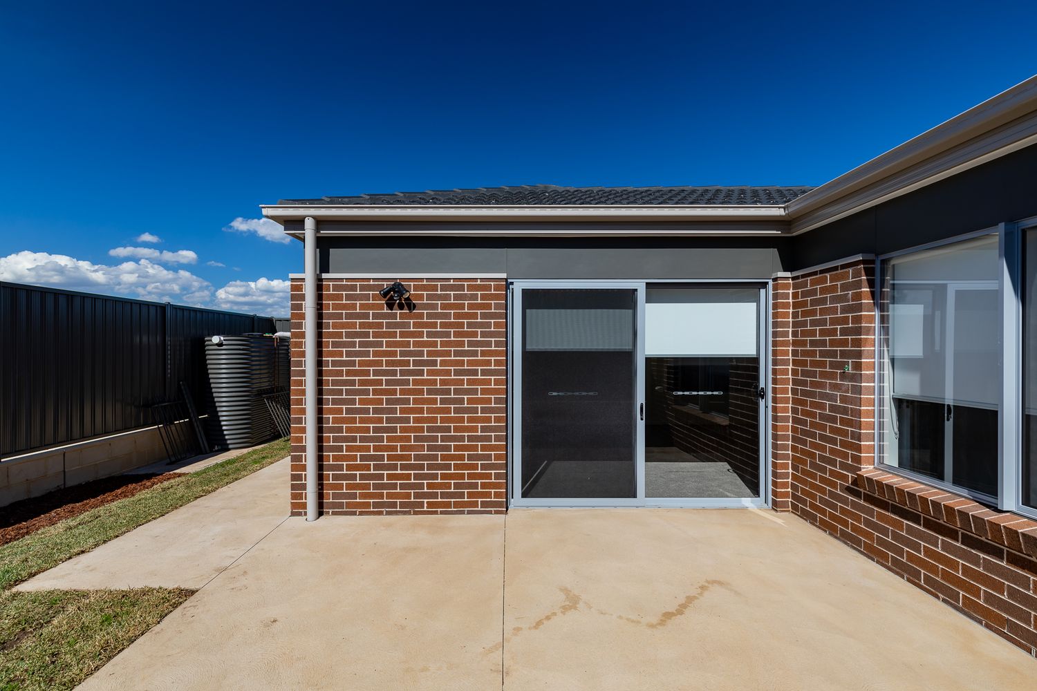 Completed brick home exterior with sliding glass doors, a concrete patio, a water tank and a dark fence under a blue sky.