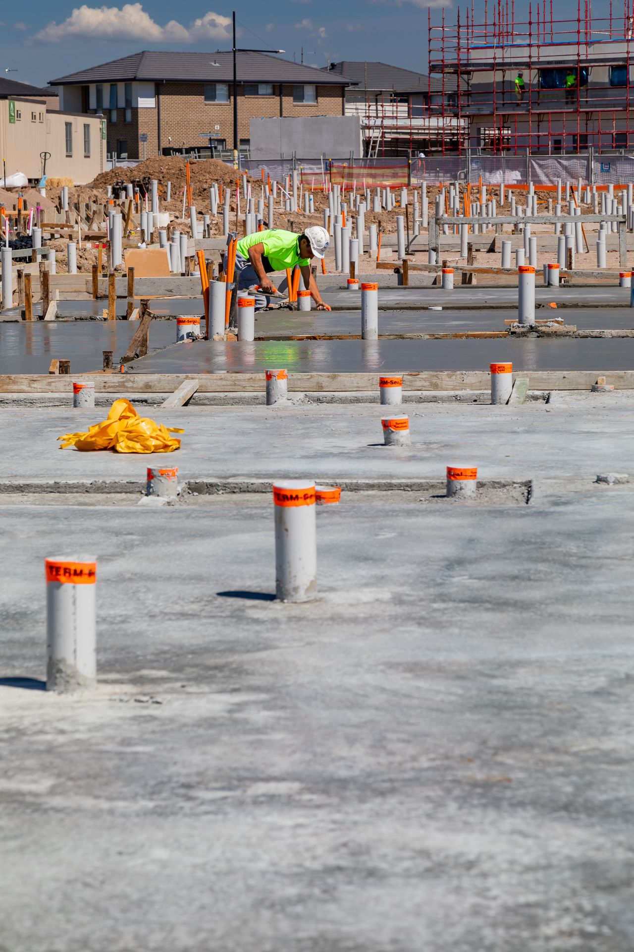 Construction worker in high-visibility gear crouches on a fresh concrete slab with capped orange pipes at a residential building site.