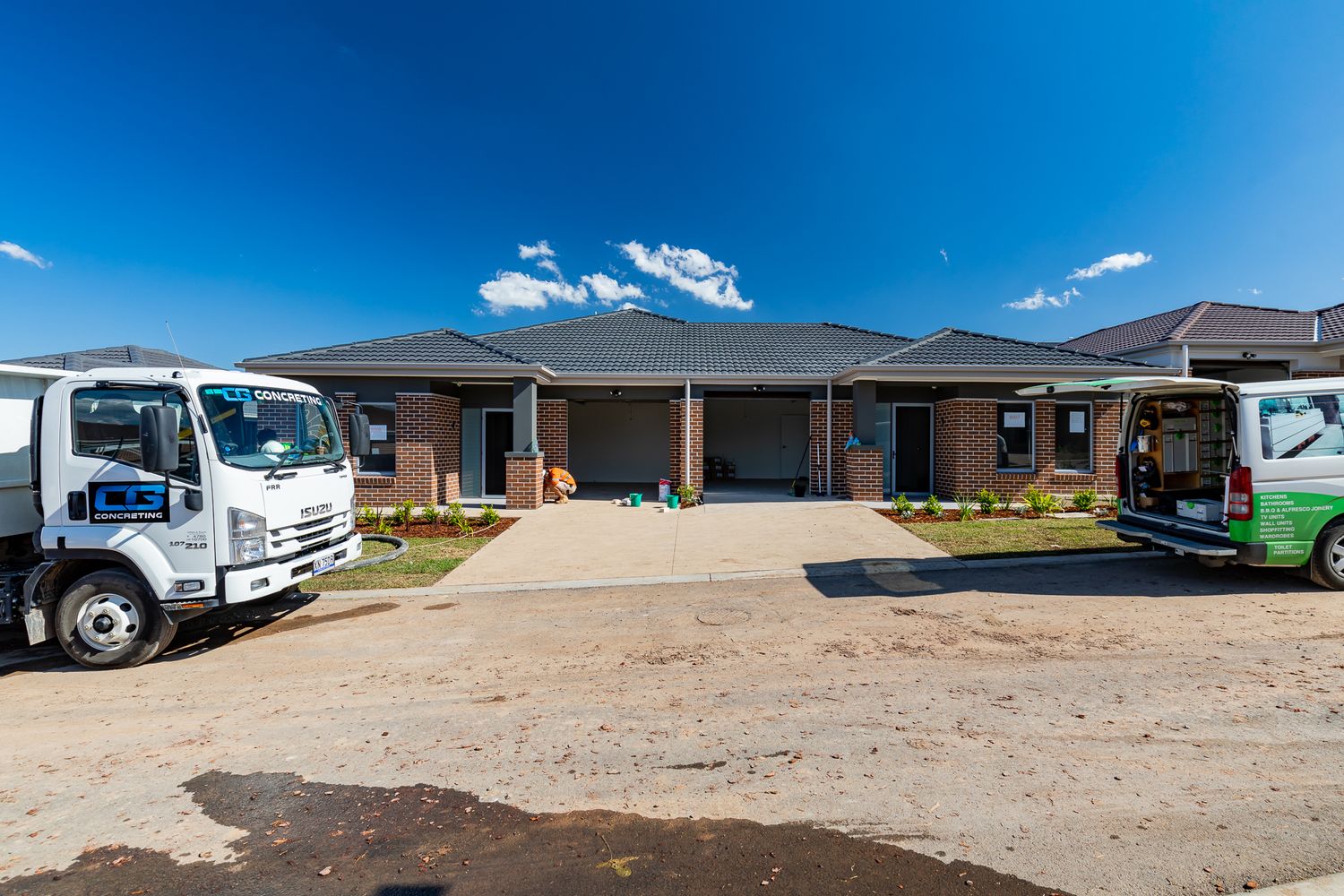 Finished brick home with a tiled roof and tidy garden, with work trucks parked on a dirt road in a new housing estate.