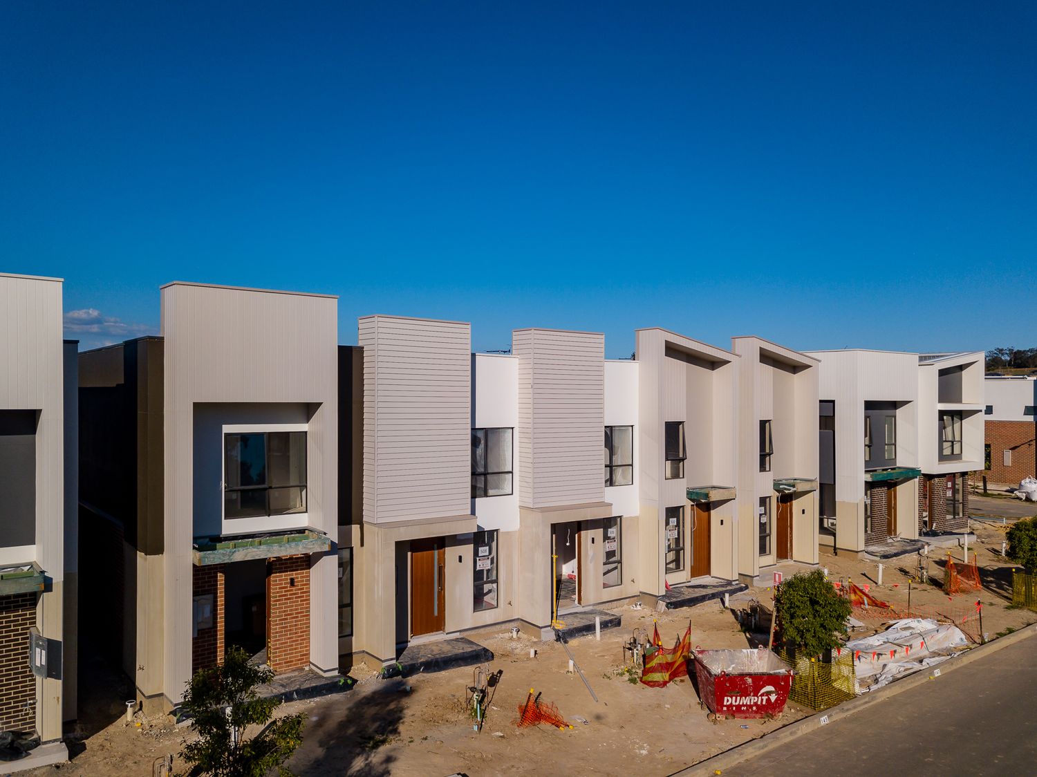 Newly built townhouses with beige cladding and timber doors beside an unfinished landscaped area under a bright blue sky.