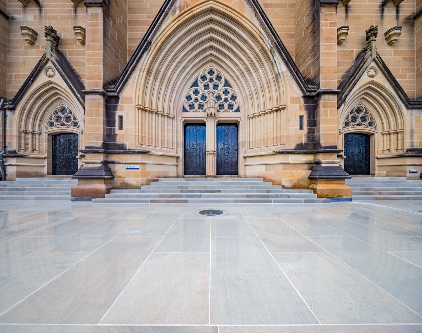 Gothic Revival sandstone cathedral facade with pointed arches, ornate rose window and dark wooden doors above a wide stone forecourt.