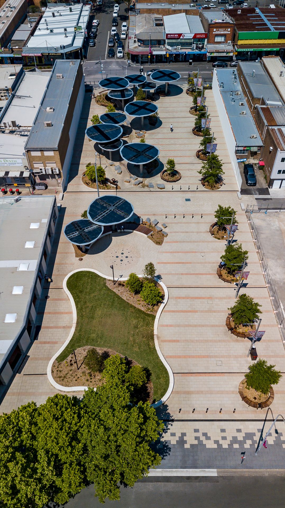 Drone view of a redeveloped town centre plaza with solar canopies, a curved lawn, young trees and neighbouring shops.
