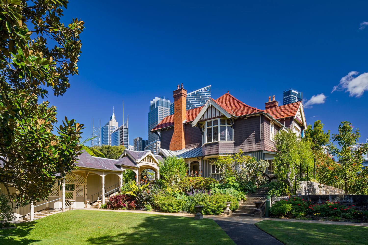 Historic Federation-style red-roofed house with a tower and lush garden, set against modern city skyscrapers under a blue sky.