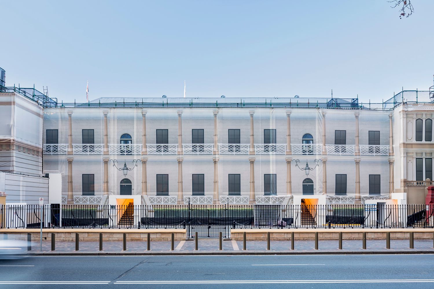 Street view of a restored neoclassical building with white balustrades, arched windows and orange entrance stairways behind a fence.