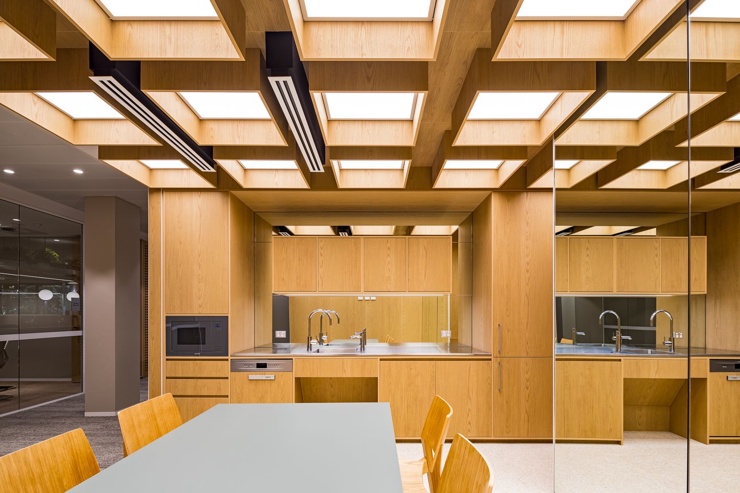 Modern office kitchenette with light oak cabinetry, exposed timber ceiling beams, linear lighting and a gray table with chairs.