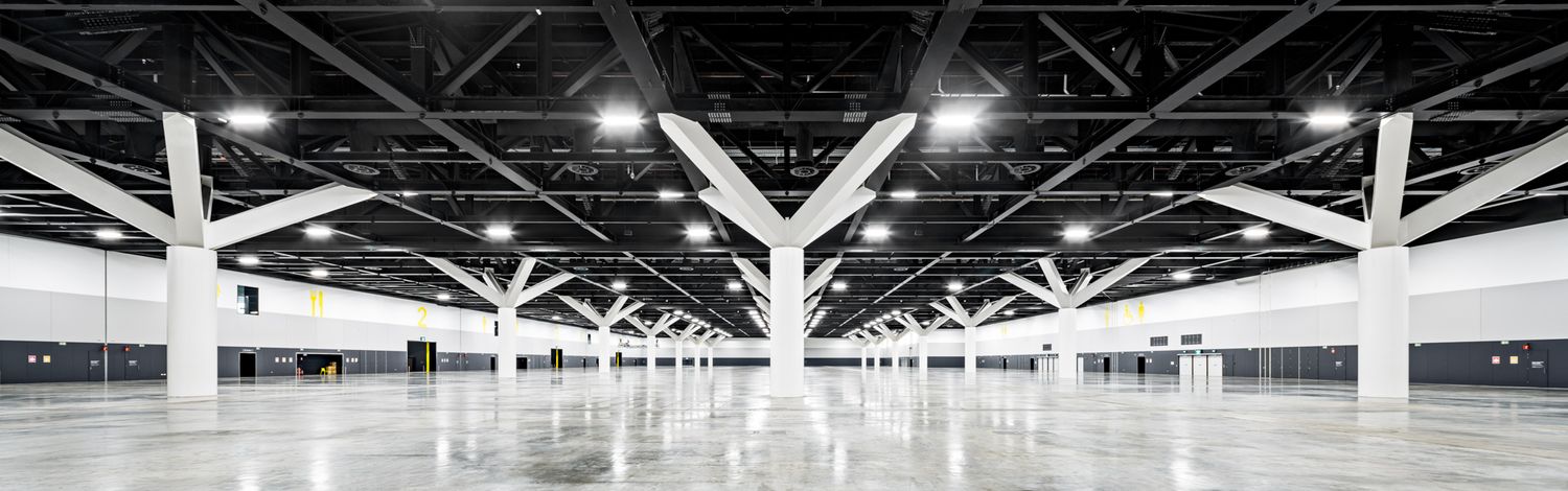 Vast exhibition hall interior with white branching columns, a polished floor and long rows of bright ceiling lights.
