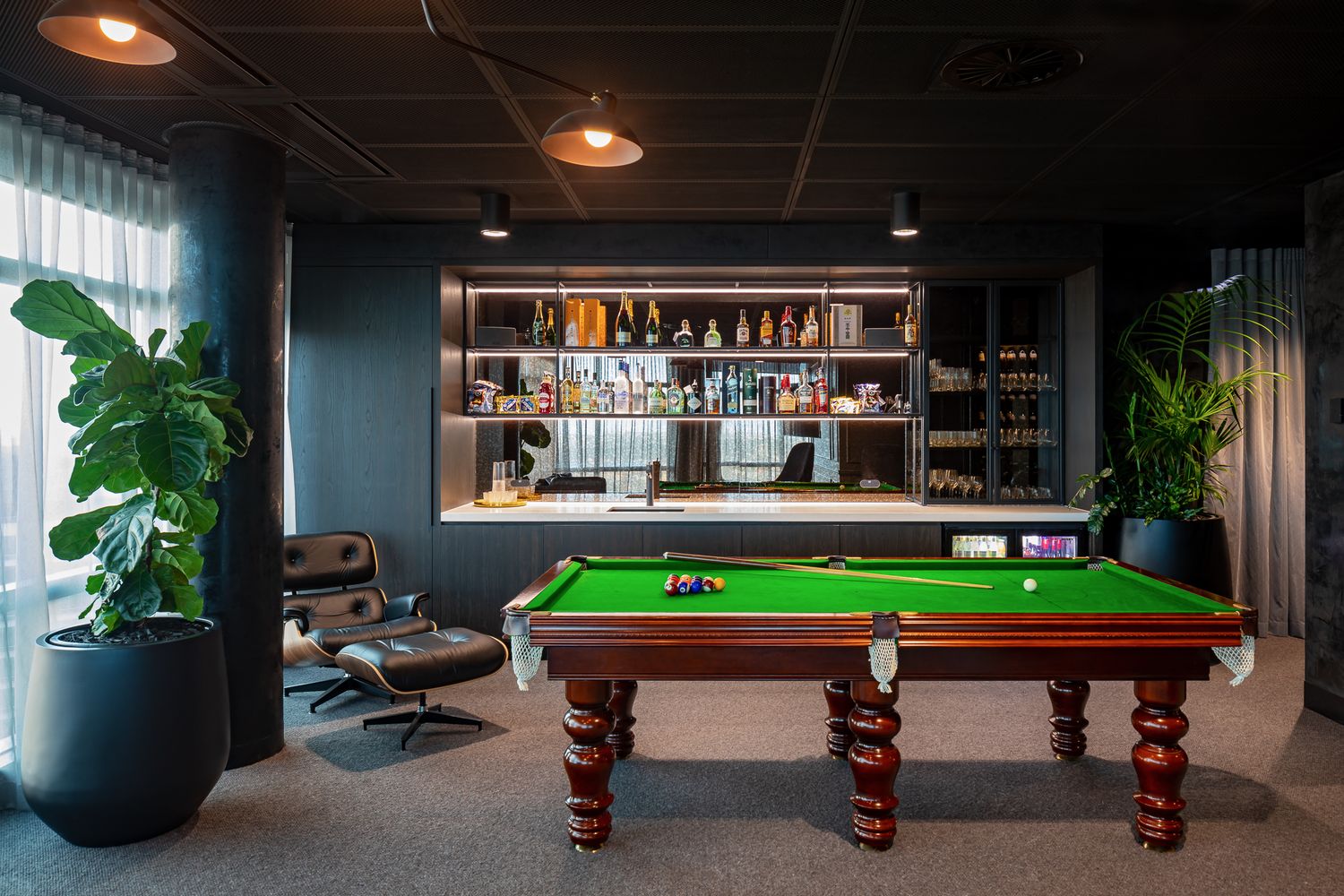 Moody games room with green billiard table, built-in bar shelving, pendant lights, leather lounge chair and large leafy indoor plants.