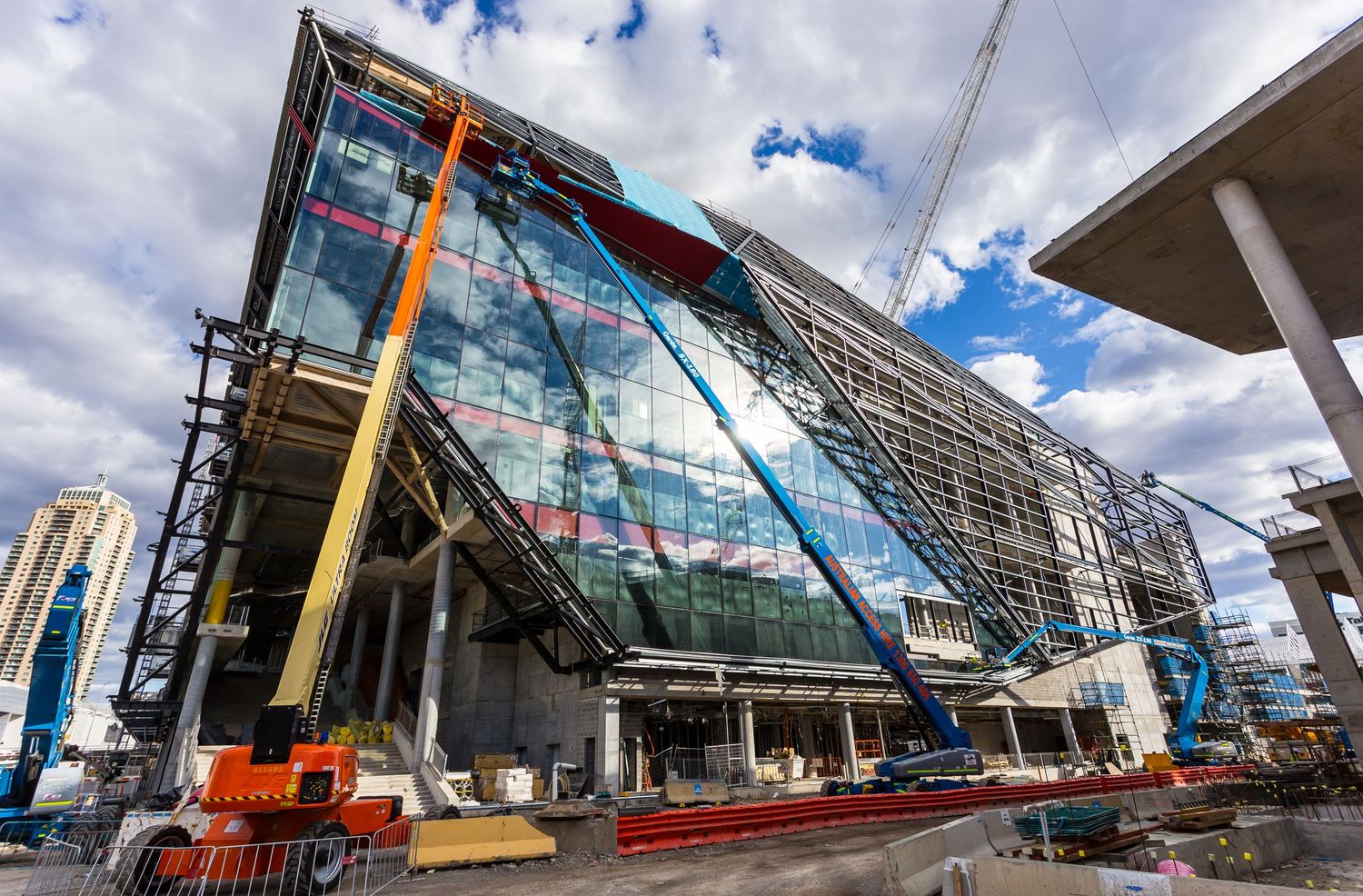 Modern glass-fronted building under construction with steel supports, scaffolding and cranes against a partly cloudy sky.