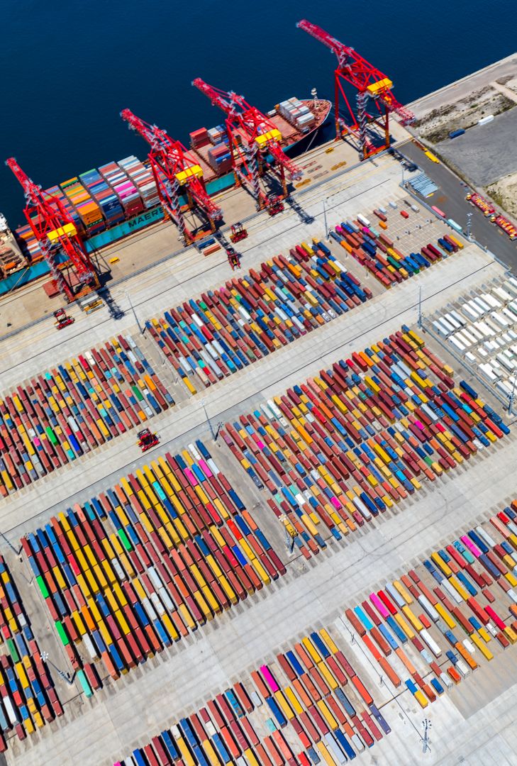 Aerial view of a busy container terminal with red gantry cranes, colorful stacked shipping containers and a cargo ship docked at the quay.