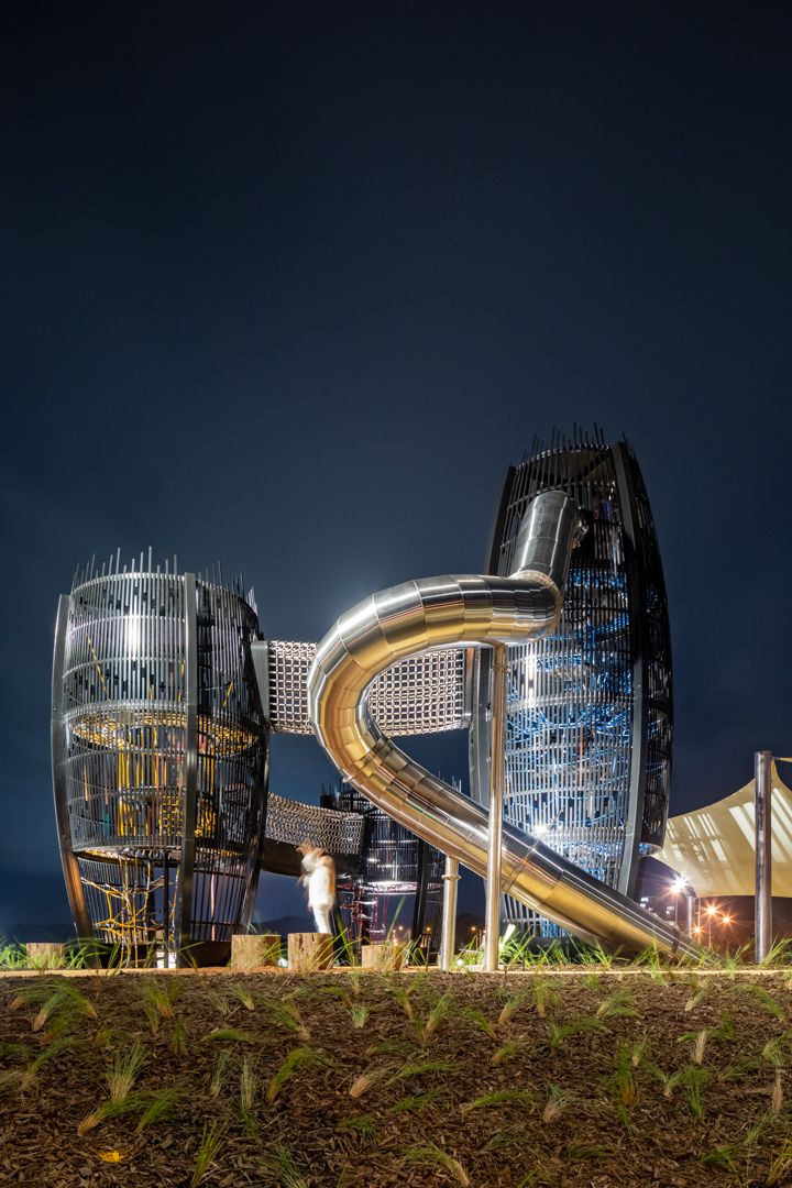 Futuristic playground at night with two mesh-clad towers connected by a curved stainless steel slide and a child standing nearby.