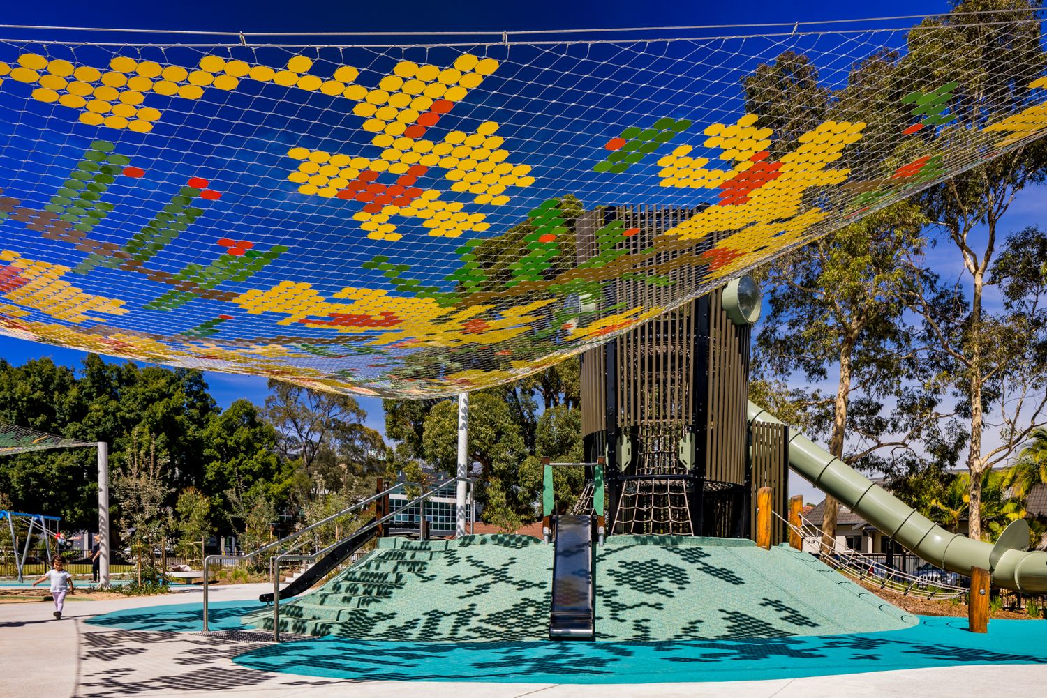 Adventure playground with a colorful rope net canopy in yellow, green and orange, a climbing tower and a long slide among trees.