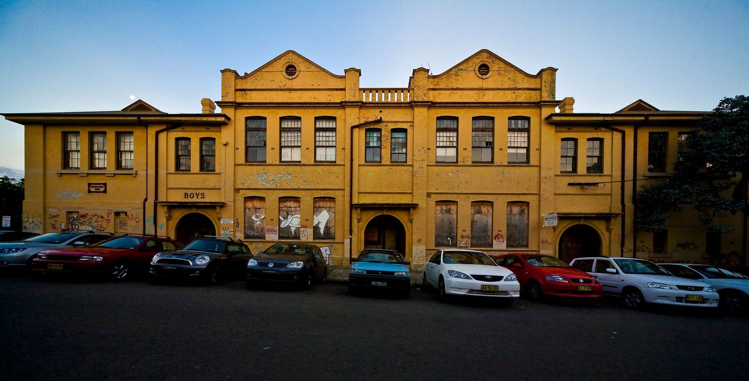 Yellow heritage building with gabled rooflines and arched doorways, with several cars parked in front at dusk.