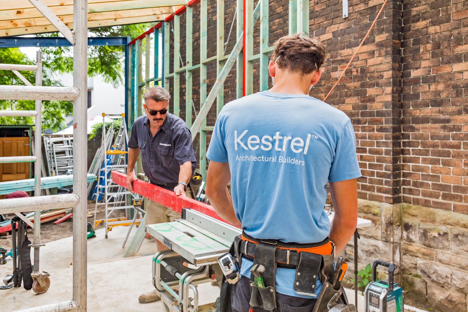 Two builders in Kestrel shirts work on a brick building renovation with scaffolding and tools nearby.