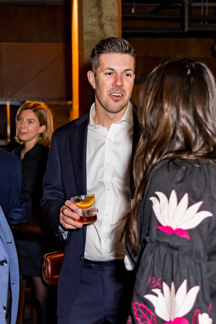 Man in a navy blazer and white shirt holds a cocktail with an orange garnish while chatting with guests at an evening networking event.