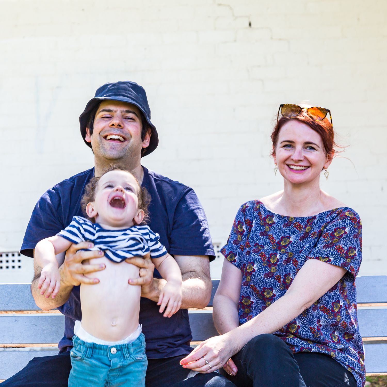 Laughing man in a bucket hat holds a giggling toddler while a smiling woman with red hair sits beside them on a bench.
