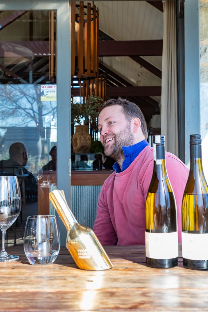 Smiling bearded man in a pink sweater sits at a wooden table with two white wine bottles, wine glasses and a gold ice scoop.