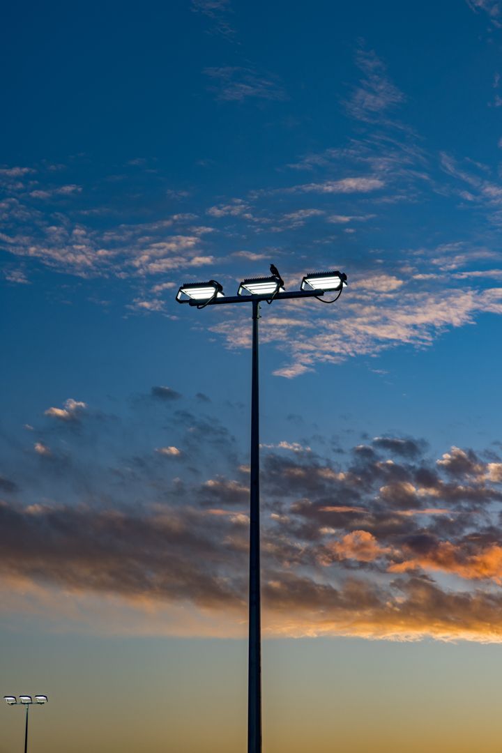Tall stadium light pole with floodlights silhouetted against a blue sky with soft clouds and an orange sunset glow.