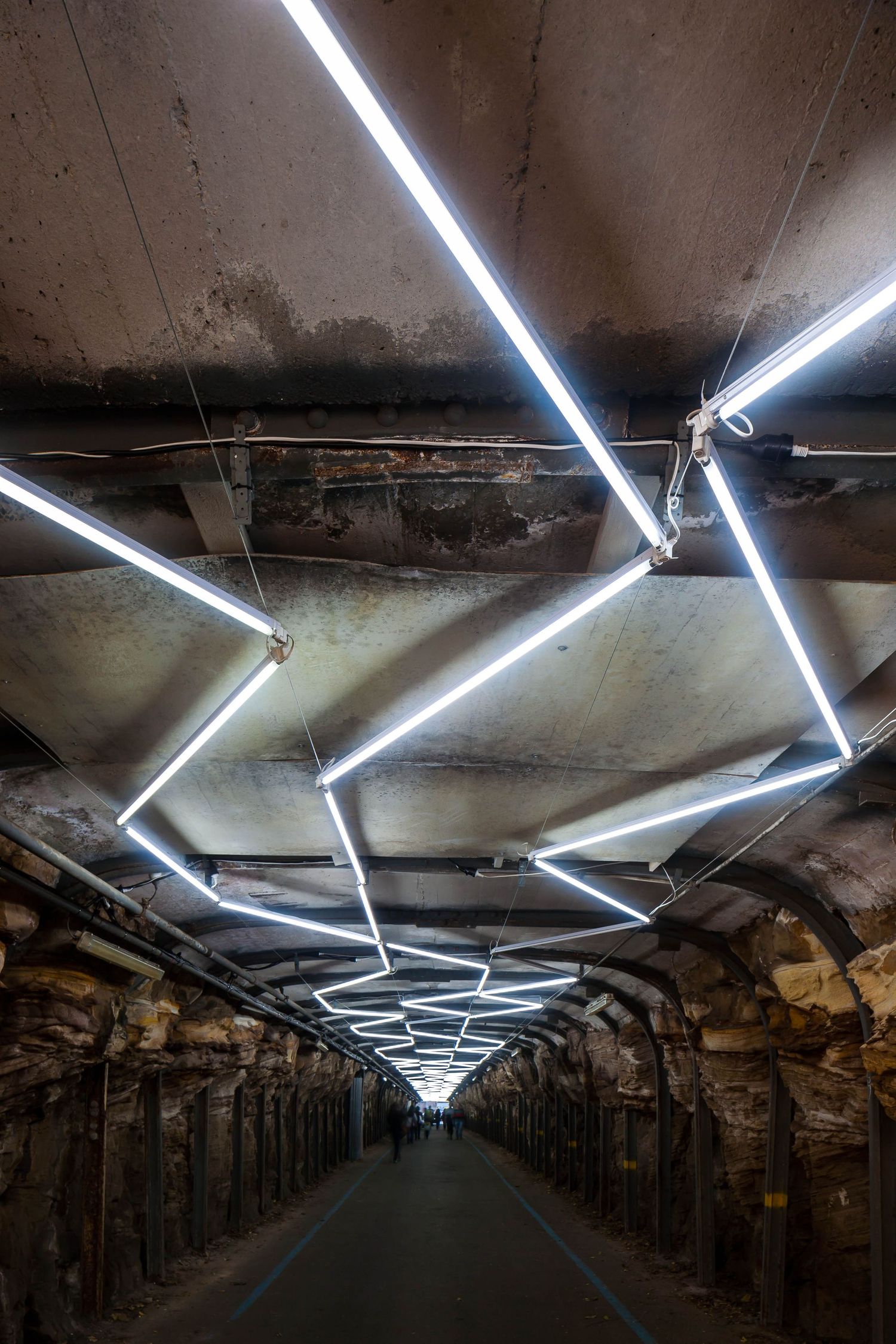 Long white tube lights are mounted in a geometric pattern on the ceiling of a historic stone tunnel with a walkway below.