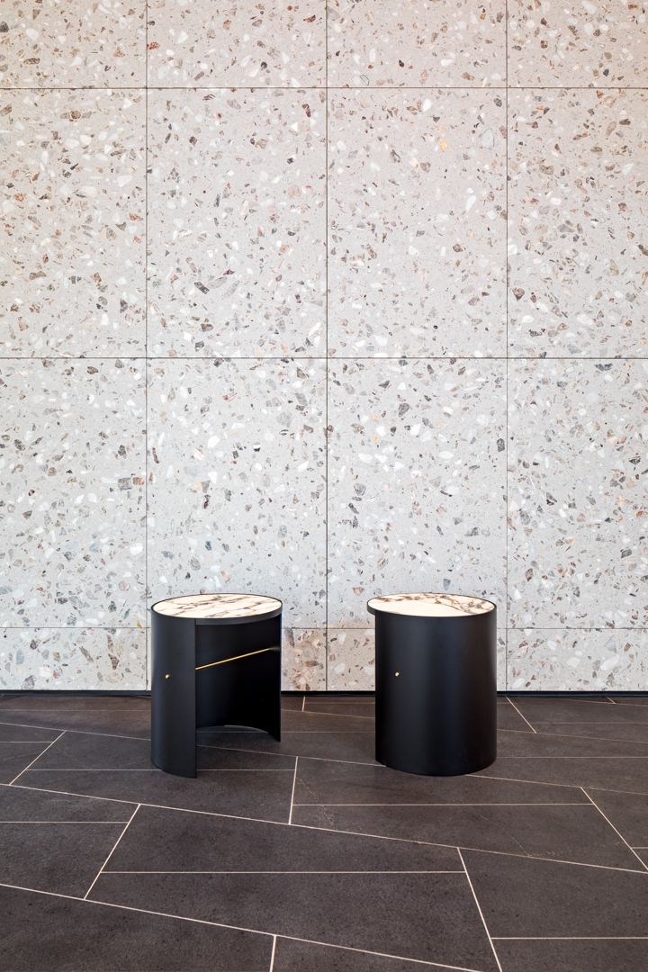 Two black cylindrical side tables with gold accents sit on dark tiled flooring in front of a speckled terrazzo feature wall.