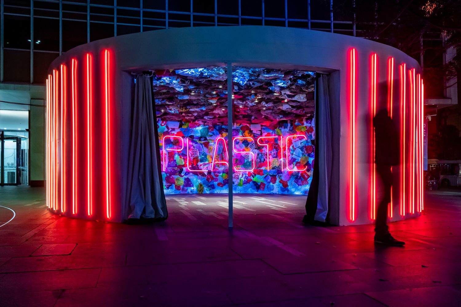 Circular pavilion with glowing red neon lights and a pink neon plastic sign over a colorful mural, with a silhouetted figure nearby.