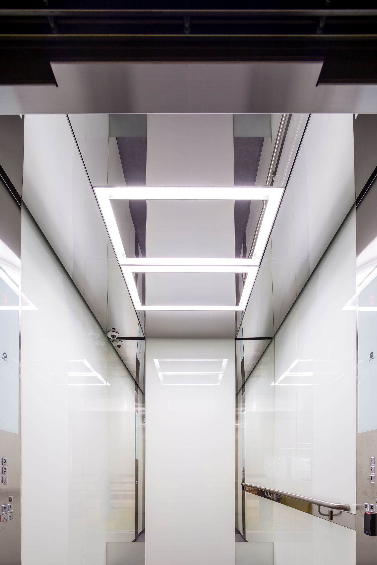 Sleek elevator interior with white glass walls, mirrored panels and a rectangular LED ceiling light fixture.