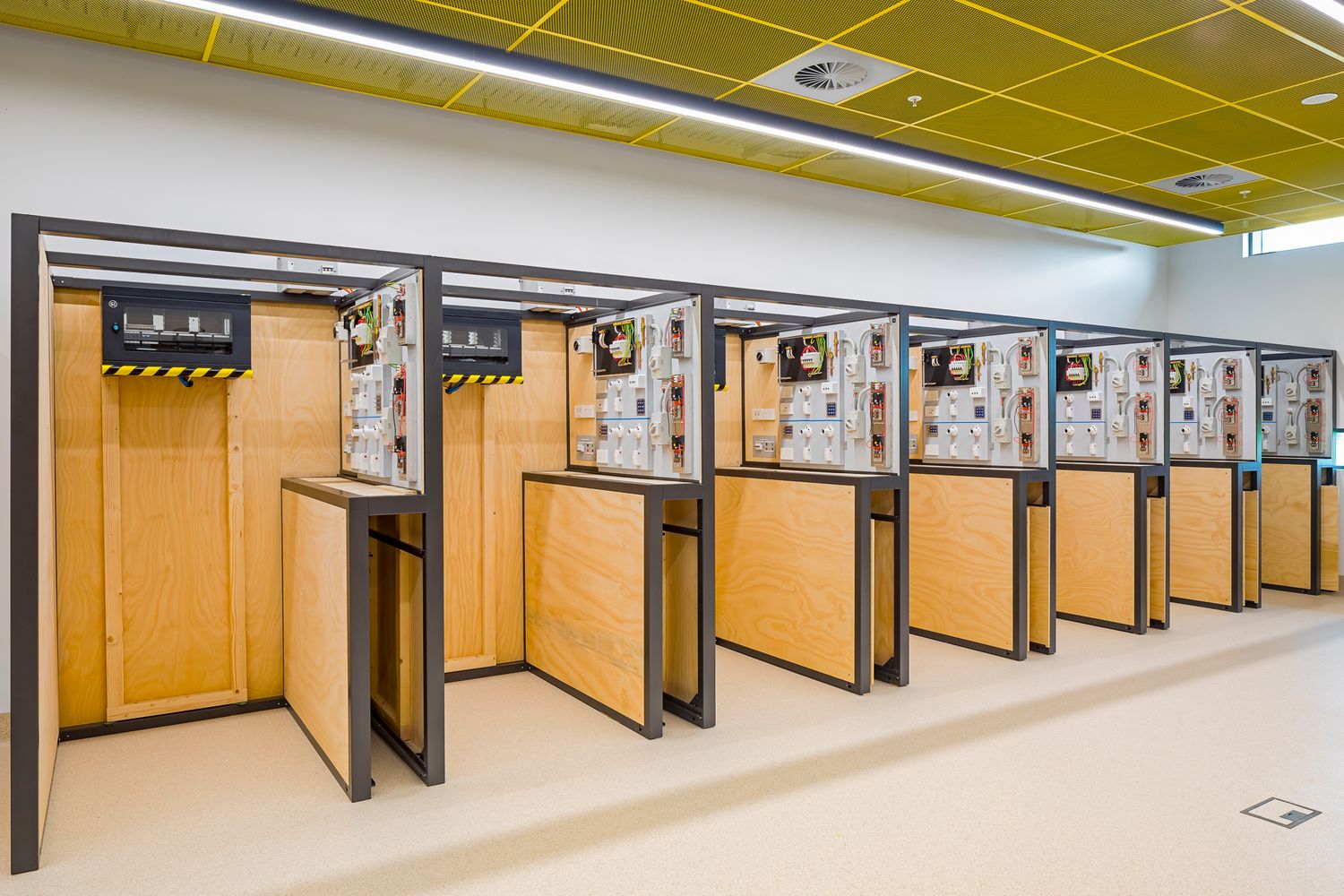 Row of timber-panelled electrical training booths with mounted equipment, set under a yellow ceiling in a bright vocational lab.