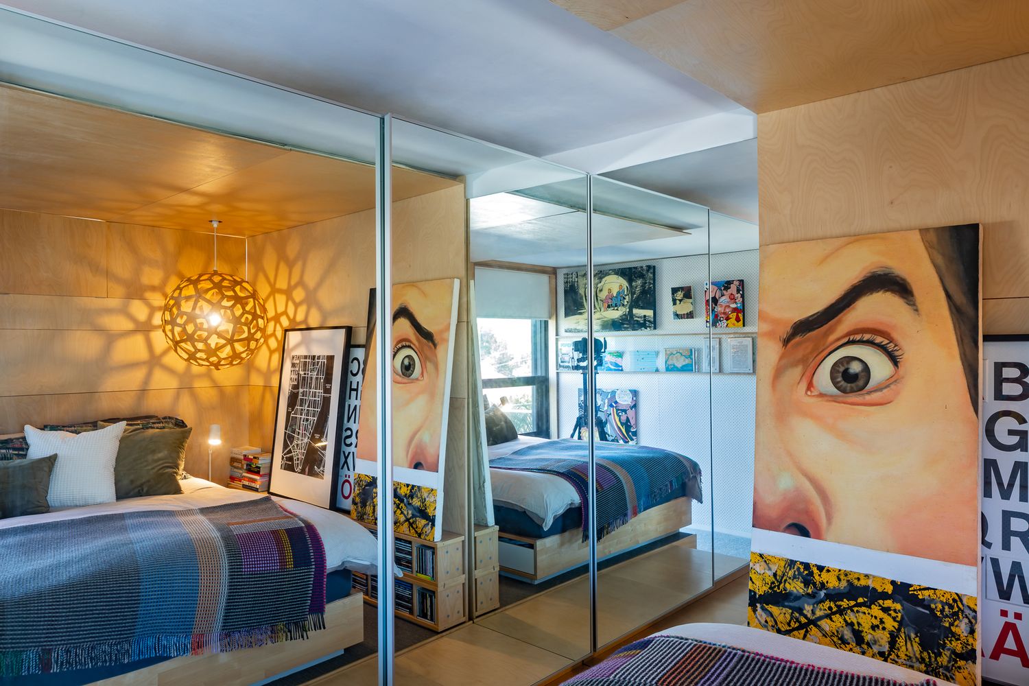 Small bedroom with mirrored wardrobe doors reflecting a patterned pendant light, bold pop-art poster, and a woven blanket.
